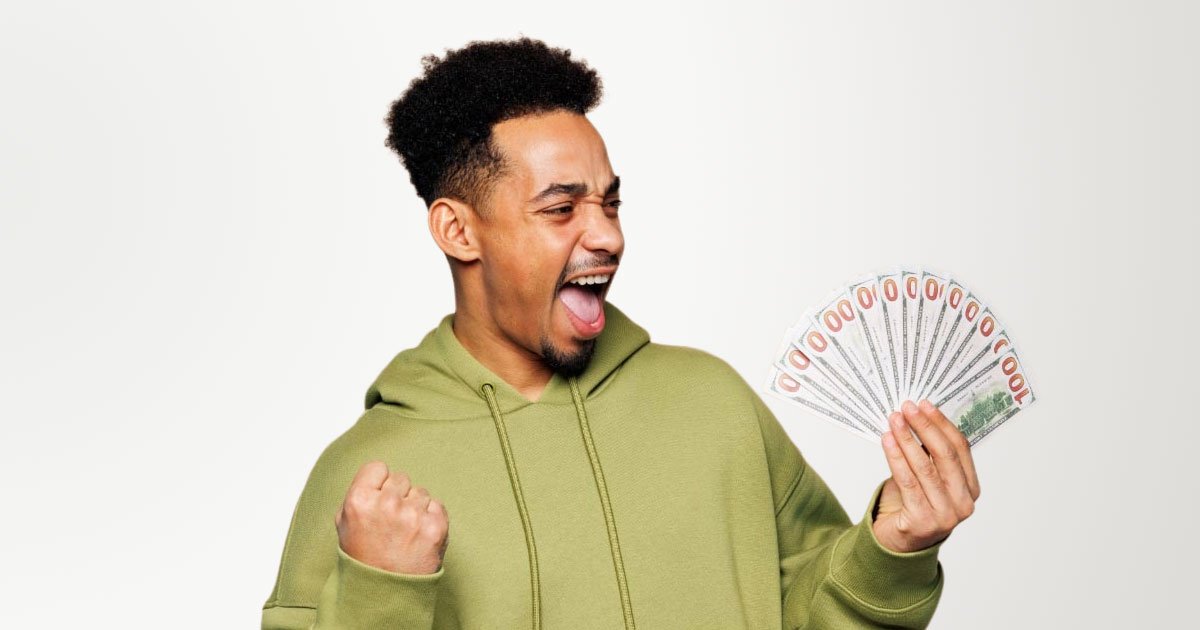African American man holding fanned out 100 dollar bills while clenching his fist wearing a green hoodie and standing in front of a beige backdrop
