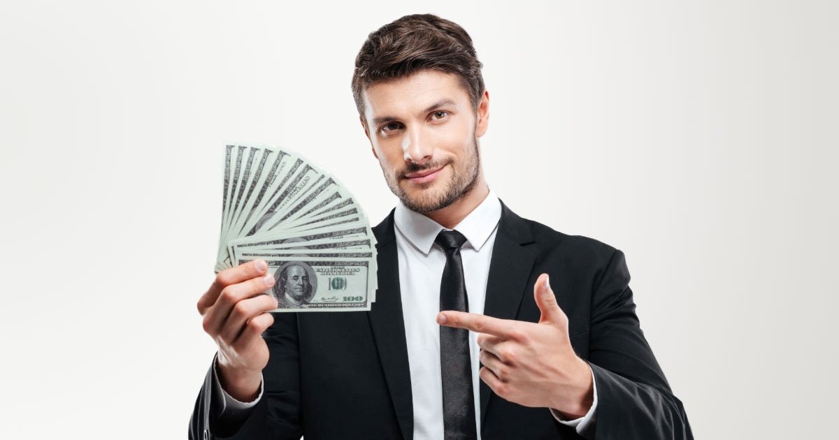 Young white guy wearing black suit and tie holding stack of cash in his right hand pointing to it with his left on a grey backing