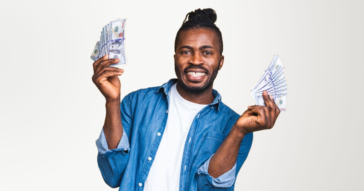 African American man with braided bun wearing denim shirt and white undershirt holding stacks of cash in each hand on a yellow backdrop