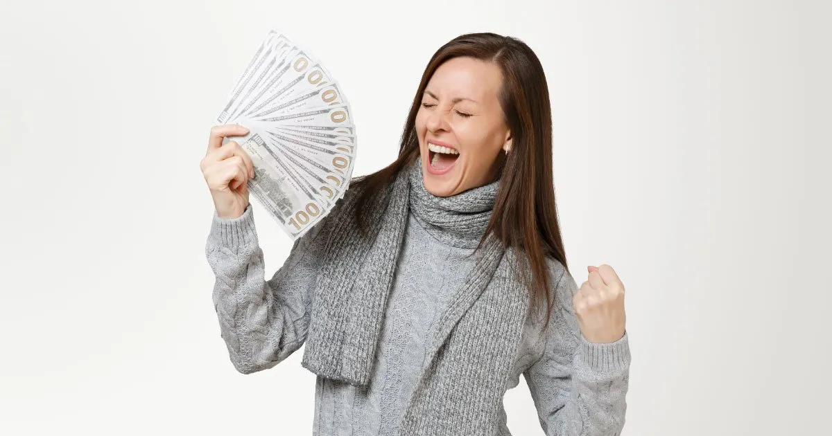 White lady with brunette straight hair wearing grey sweater and scarf celebrating the cash she's holding in her right hand