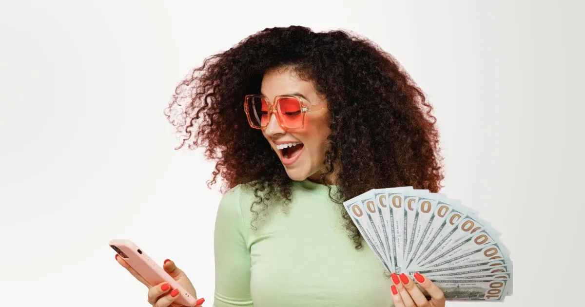 African American woman laughing while looking at her phone in her right hand and holding stack of cash in her left with a pin background
