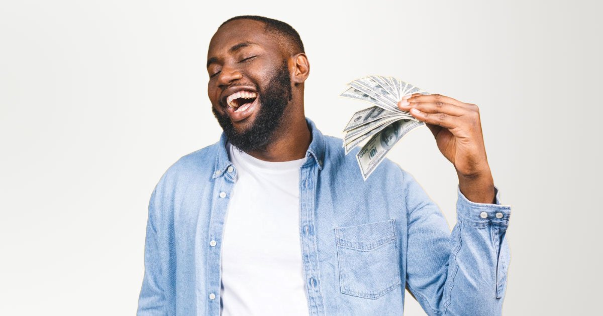 Young black man wearing unbuttoned denim shirt with white undershirt fanning himself with a stack of 100s on yellow backdrop