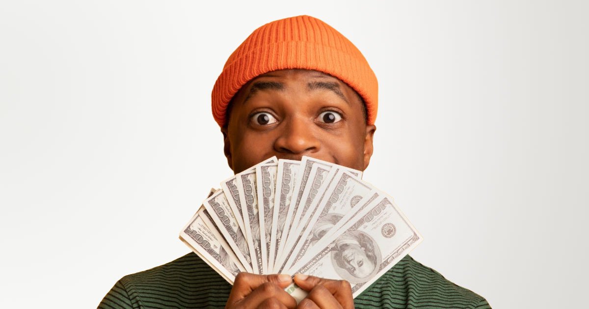 Black guy wearing reddish-orange beanie and green shirt with black stripes hiding his mouth behind a fanned out stack of 100s in front of yellow background