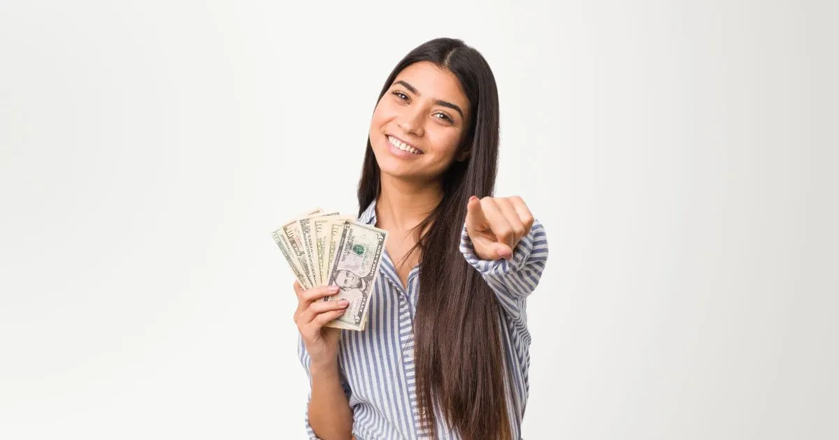 Brunette woman wearing striped button shirt with sleeves rolled up holding cash pointing at camera man on white background