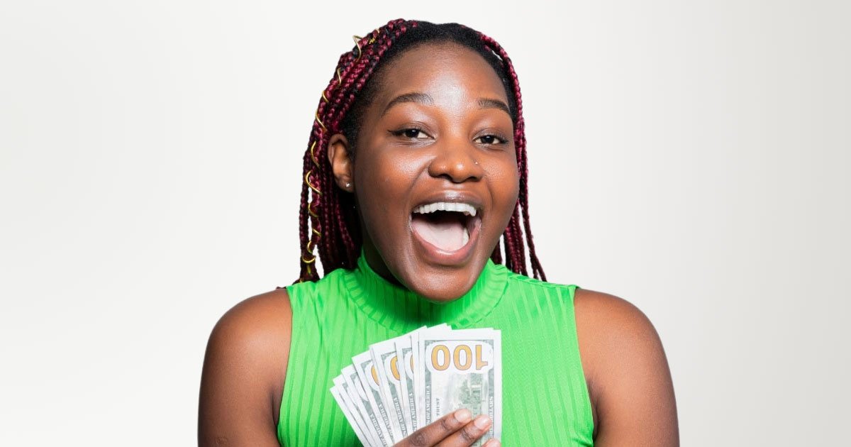Black woman with pink braids excitedly holding hundred dollar bills wearing a green sleeveless shirt with a grey background
