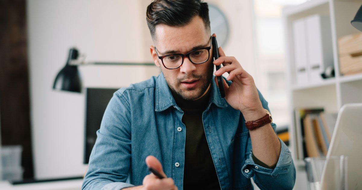 White guy wearing denim button down shirt and glasses on his mobile phone and taking notes at his desk