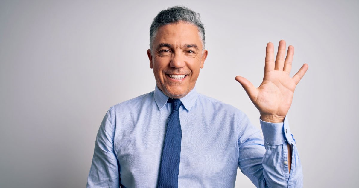 Caucasian man wearing blue long-sleeve with dark blue tie holding hand up to say hi while smiling
