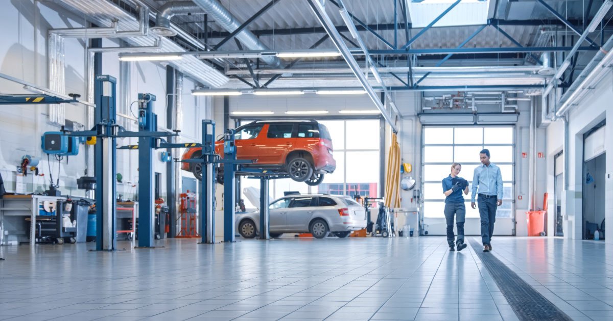 Mechanic walking through shop with her customer going over repair details with an orange car on a lift and silver station wagon in the background